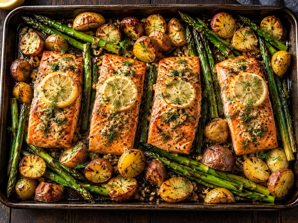 Four roasted salmon fillets with lemon slices and dill on a sheet pan with asparagus and baby potatoes